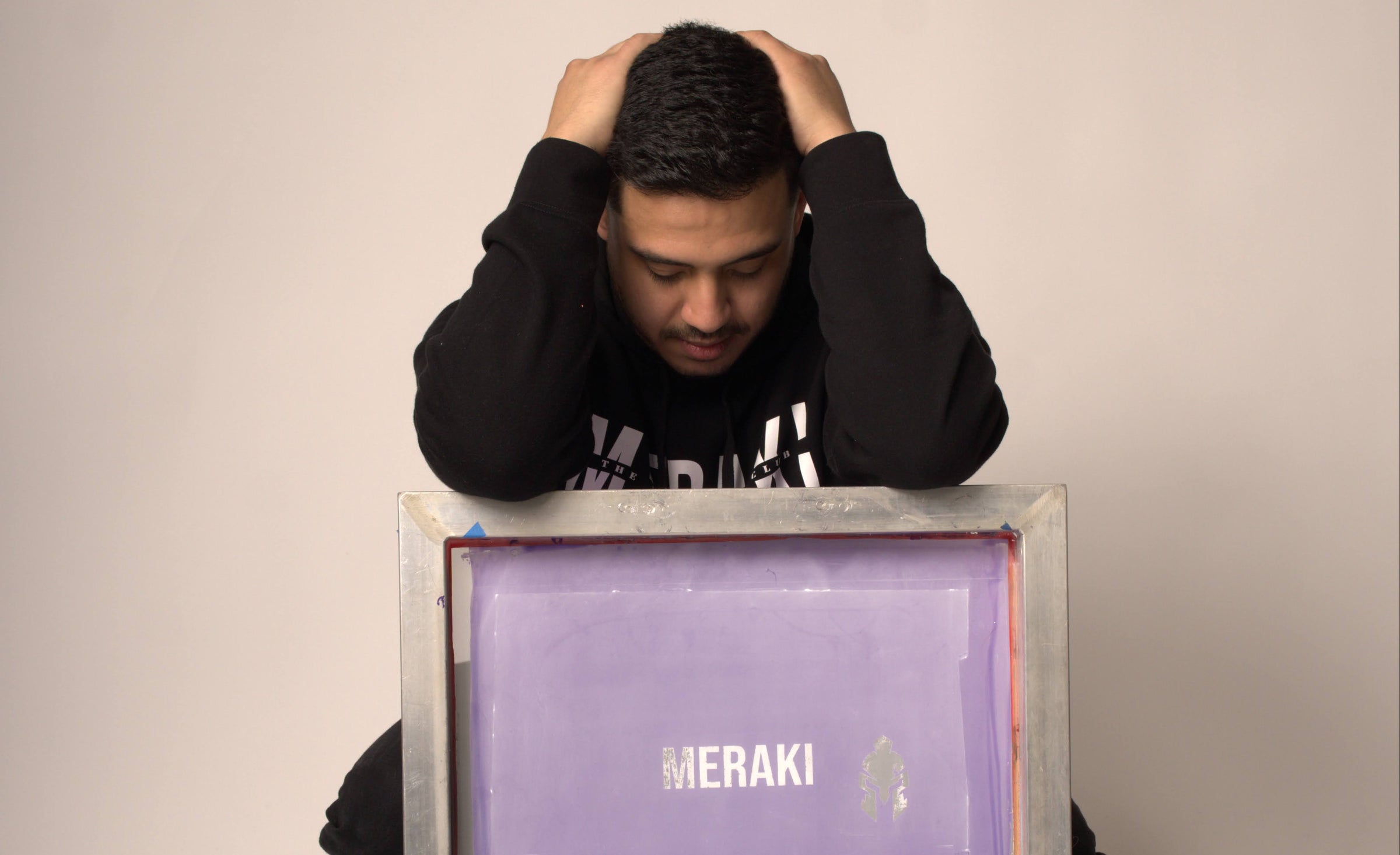 Louie Martinez crouches behind a purple screen-printing frame labeled MERAKI, symbolizing the brand’s garage origins and hands-on craft.